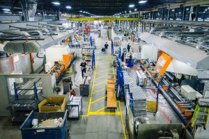 EPSYLON - Vue intérieure d'une usine moderne avec des travailleurs opérant des machines industrielles sur une ligne de production, mettant en avant l'efficacité industrielle.